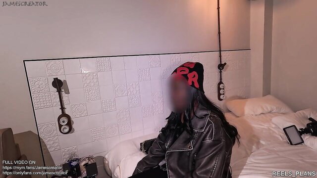 Hot Asian girl in leather jacket and red beanie sits on bed, blurred face