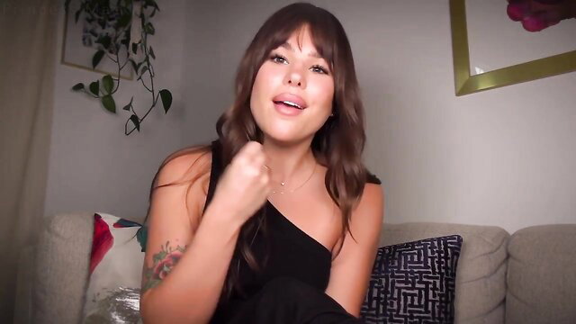 Brunette with bangs smiling in one-shoulder black dress, tattooed arm