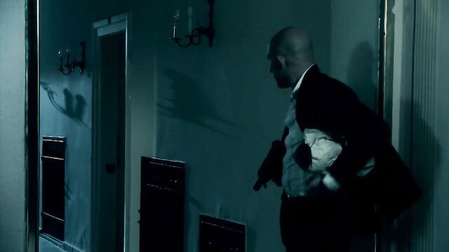 Bald suited man in dim room holds teddy bear and gun in thriller porn scene