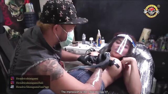 Asian tattoo artist inks topless woman's breast in studio closeup