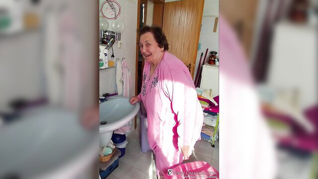Smiling mature granny in pink robe at sink, big melons tease