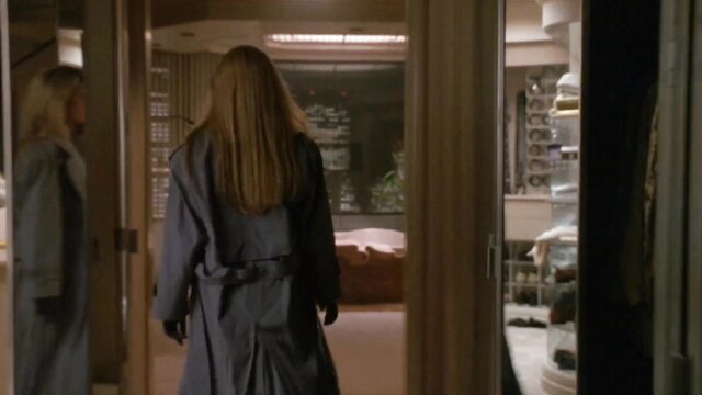 Kim Basinger in a gray trench coat walking into a bedroom