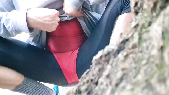 Transgender in spandex swimsuit pulling aside red crotch outdoors by tree