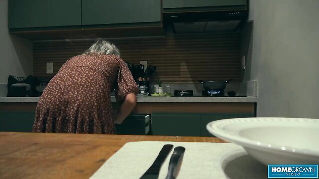 Elderly gray-haired amateur in floral dress bends over kitchen counter