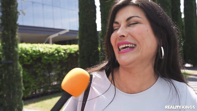 Audrey Reid smiling with mic in outdoor interview, hot brunette MILF