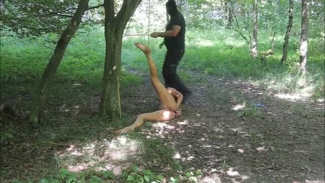 Naked woman bound and suspended in forest by masked man in outdoor BDSM scene