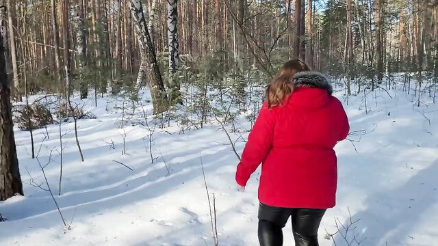 Curvy redhead in red parka and tight leather pants walks in snowy forest