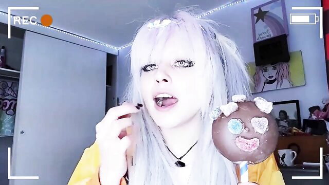 Pale goth girl with white hair sticks out tongue holding clown lollipop