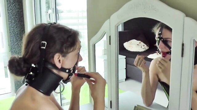 BDSM sub brunette applying lipstick in leather collar harness gag mirror