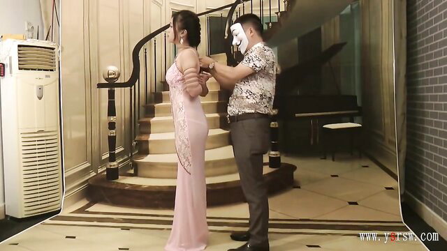 Chinese girl in pink long dress bound wrists by man in Guy Fawkes mask BDSM