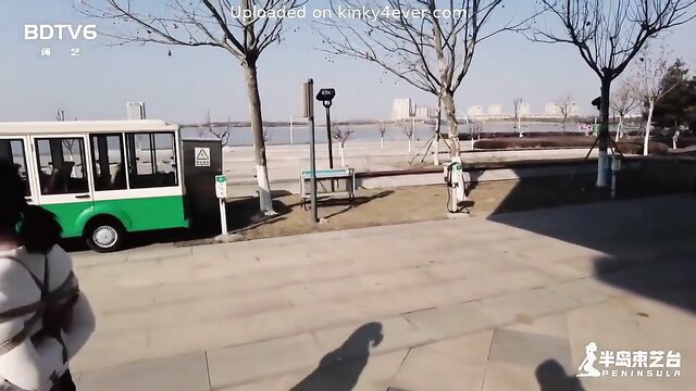 Chinese woman bound outdoors in white straps near green bus BDSM tease