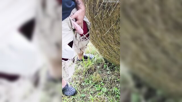 Man in khaki shorts and Crocs pulls out cock peeing by hay bale outdoors