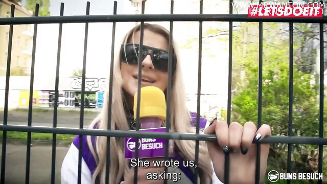 Blonde teen Kylie Kay in sunglasses and varsity jacket behind fence, sucking bottle