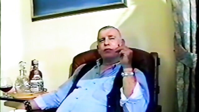 Bald mature man in vest smokes and sips wine in BDSM lounge