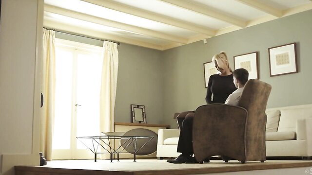 Blonde woman in black dress stands over seated man in elegant SexArt scene