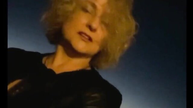 Blonde MILF in black leather top, seductive gaze, adult video thumbnail