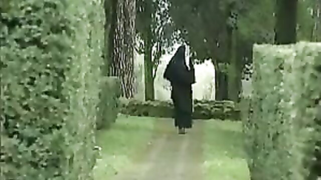 Bushy nun in black habit walks through green garden hedges, unshaved hairy tease