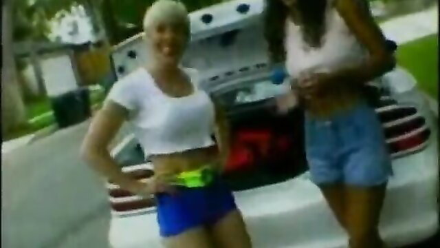 Blonde and black women in shorts posing by car trunk in hot outdoor scene