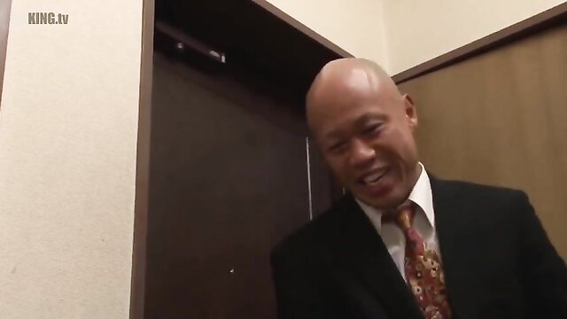 Bald Asian man in suit smiling slyly at door in JAV