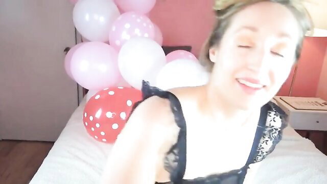 Blonde in black lace lingerie surrounded by pink balloons on bed