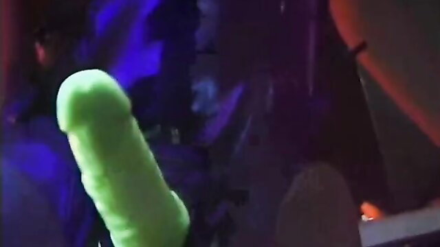 Pirate woman thrusts glowing green strap-on dildo in neon-lit fetish scene