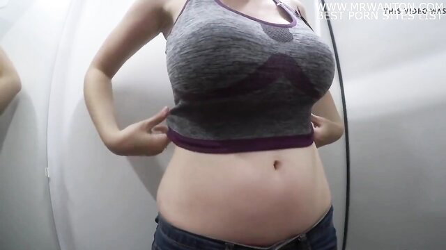 Bianca lifts gray sports bra in fitting room, showing pregnant belly
