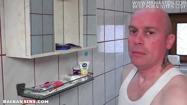 Bald tattooed Balkan man in white tank top stares in bathroom selfie