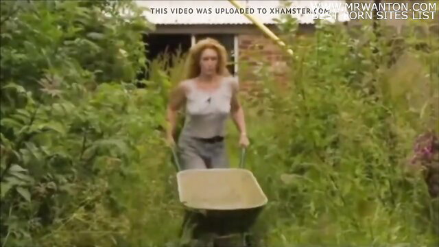 Charlie Dimmock pushing wheelbarrow in garden, braless in tank top
