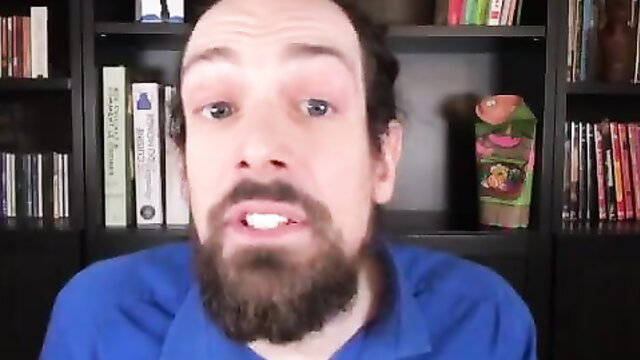Bearded man with long hair smirks in blue shirt, bookshelf background