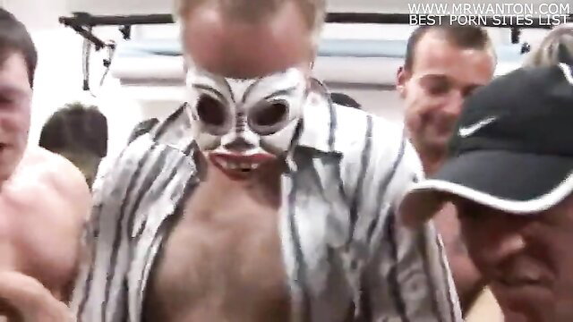 Bald masked man in open shirt surrounded by nude men in gym gangbang