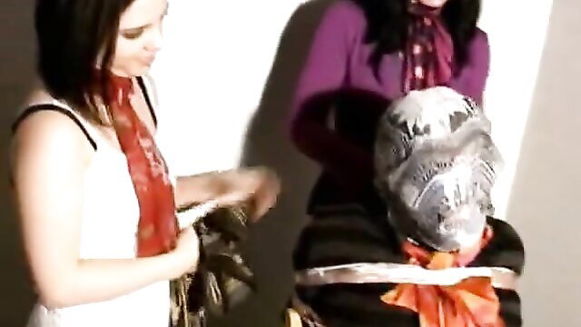 Two women mummify bound man in tape and scarves BDSM bondage