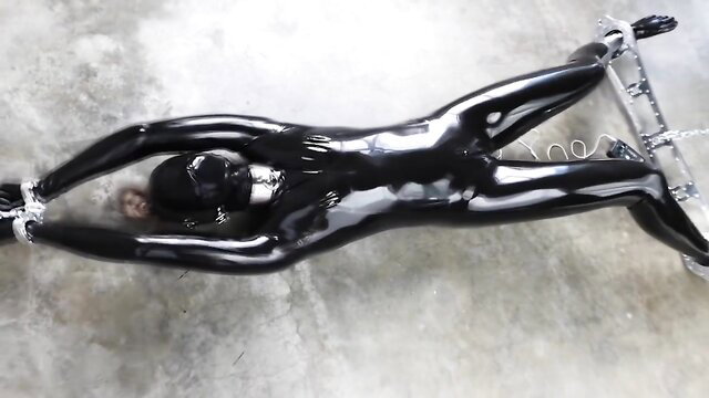 Rubber-bound girl in latex restrained on metal frame for electro BDSM play