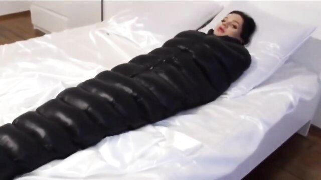 Brunette babe zipped tight in black latex sleepsack bondage on bed