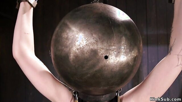 Charlotte Sartre locked in metal bondage ball, arms raised, pale skin tattooed
