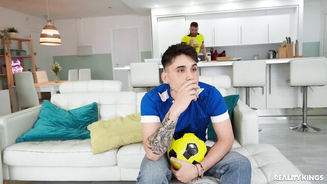 Xander Corvus in blue soccer jersey holds ball on couch, Lena Coxx in kitchen