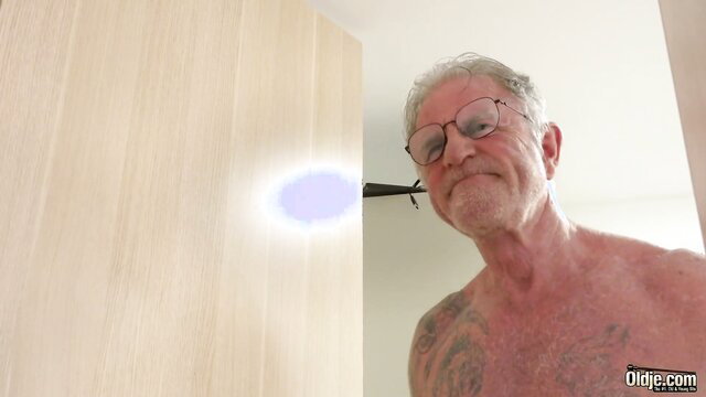 Old man with glasses and tattoos smiles shirtless near Bella Sinn