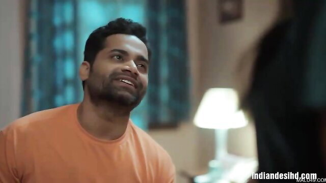 Handsome Indian man in orange tee smirks intimately in bedroom scene