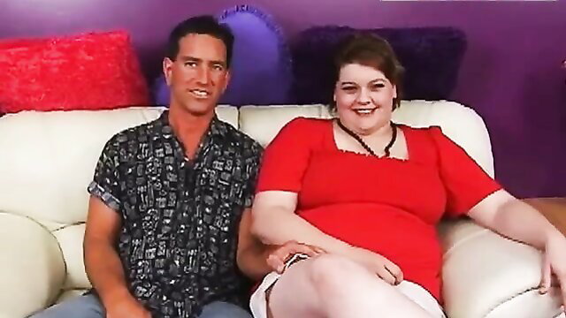 Curvy redhead BBW in red dress smiles with man on couch in horny preview