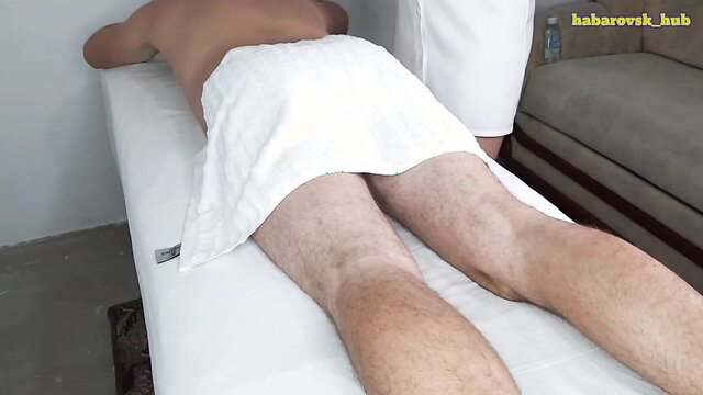 Asian masseuse in white uniform kneels behind nude male client on table