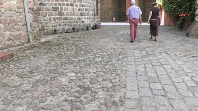 Older couple walks hand-in-hand on cobblestone street near stone wall
