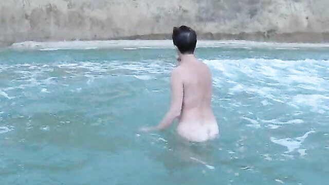 Nude man with short black hair standing naked in turquoise ocean water from behind