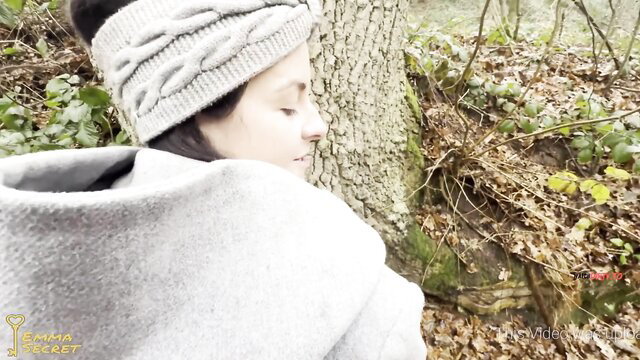 EmmaSecret in cozy gray headband and poncho, leaning on tree in forest