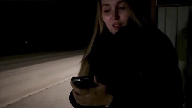 Blonde Russian stepmom in coat checks phone at night bus stop