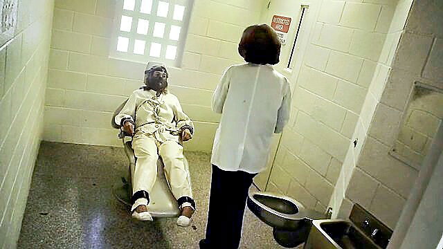 Black prisoner restrained in white jumpsuit, female doctor in cell