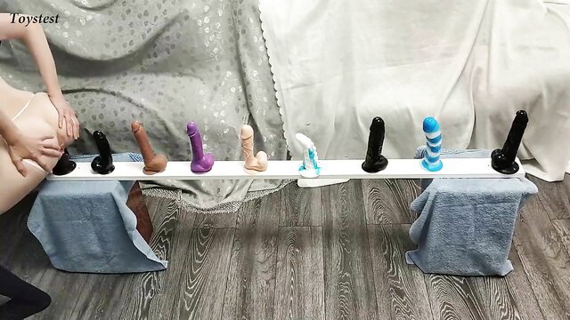 Tiny babe tests row of colorful dildos on table, nude ass up
