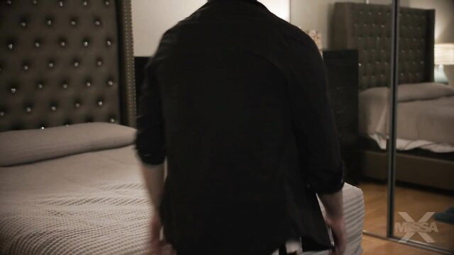 Man in black shirt approaches bed in dimly lit bedroom scene