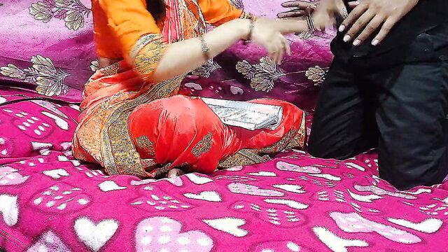 Indian teacher in red saree sits on bed with man kneeling, exam paper prop