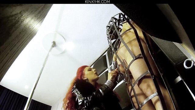 Redhead dominatrix in leather straps nude male slave in metal bondage cage
