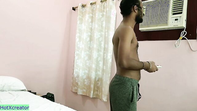 Shirtless Indian man in grey sweats holding game controller in hotel room