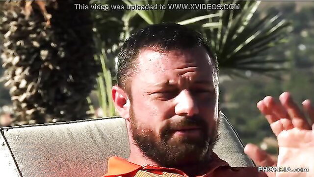 Bearded man in orange shirt waves hand outdoors, gay teasing thumbnail